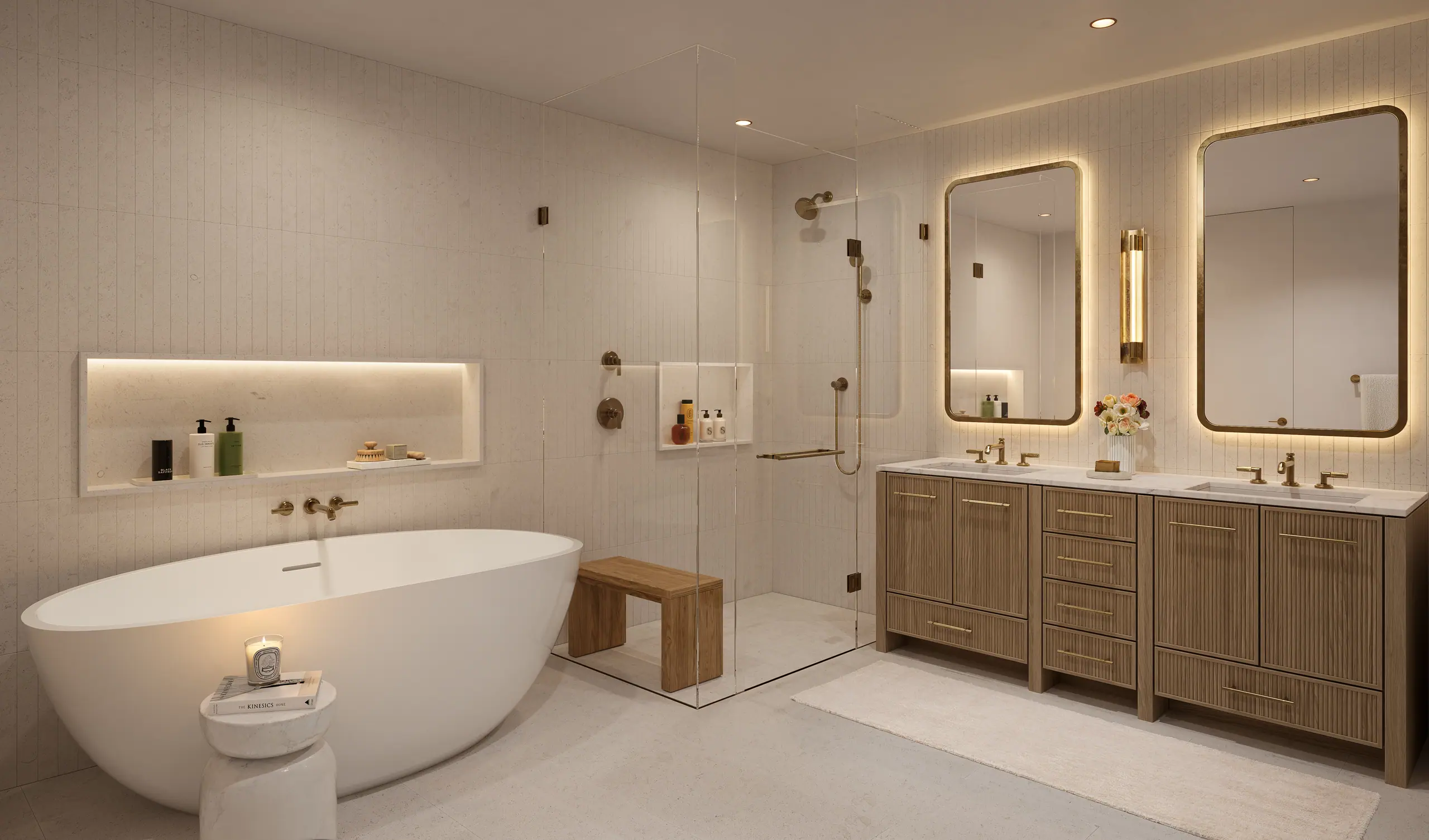Primary bathroom in pale stone with a freestanding oval tub, a glass-enclosed walk-in shower with brass fittings, an oak double vanity and a backlit rounded mirror.