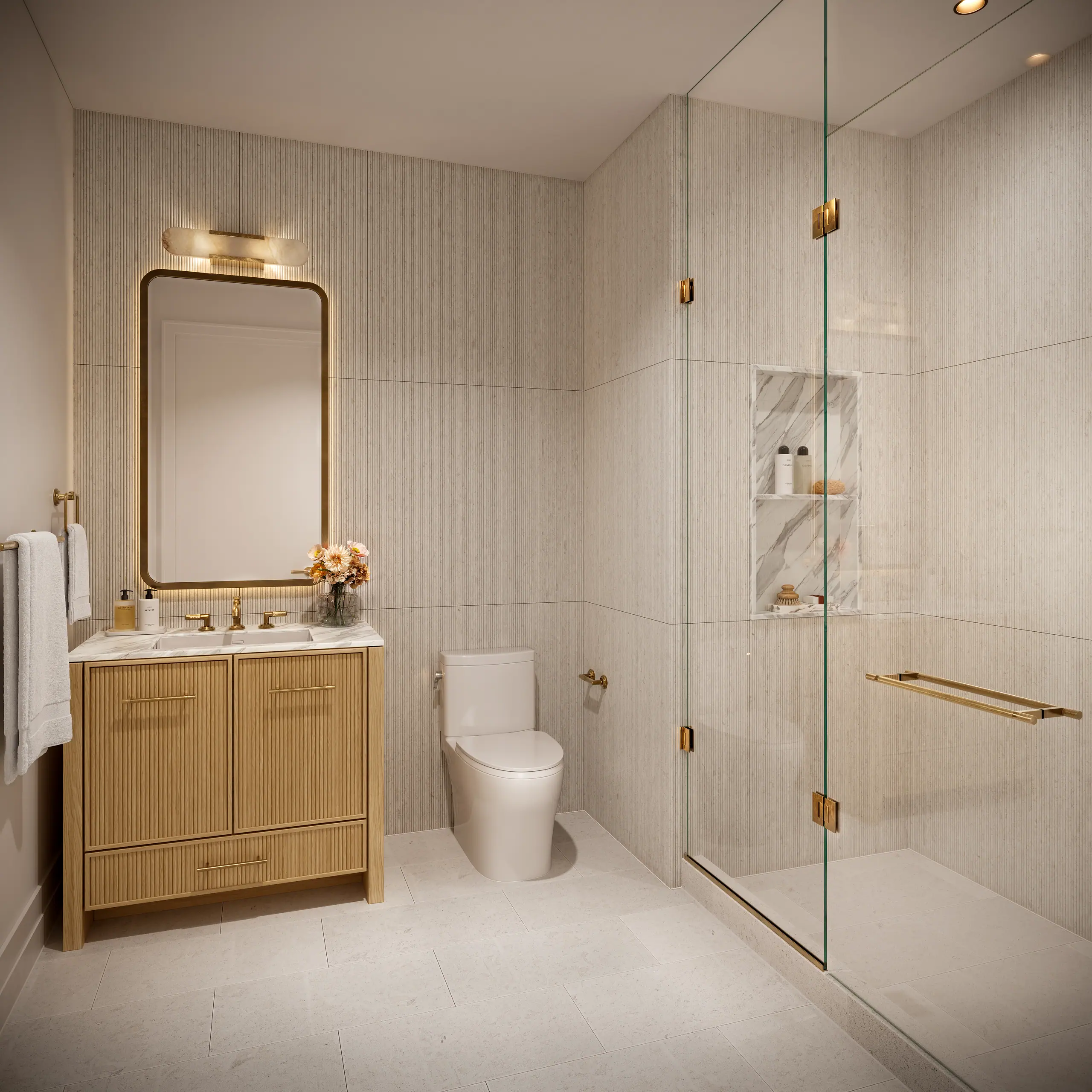 Bathroom with an oak vanity and brass hardware, a rounded mirror under a brass wall light, a toilet, and a glass-enclosed shower with brass fittings against pale vertical tiles.